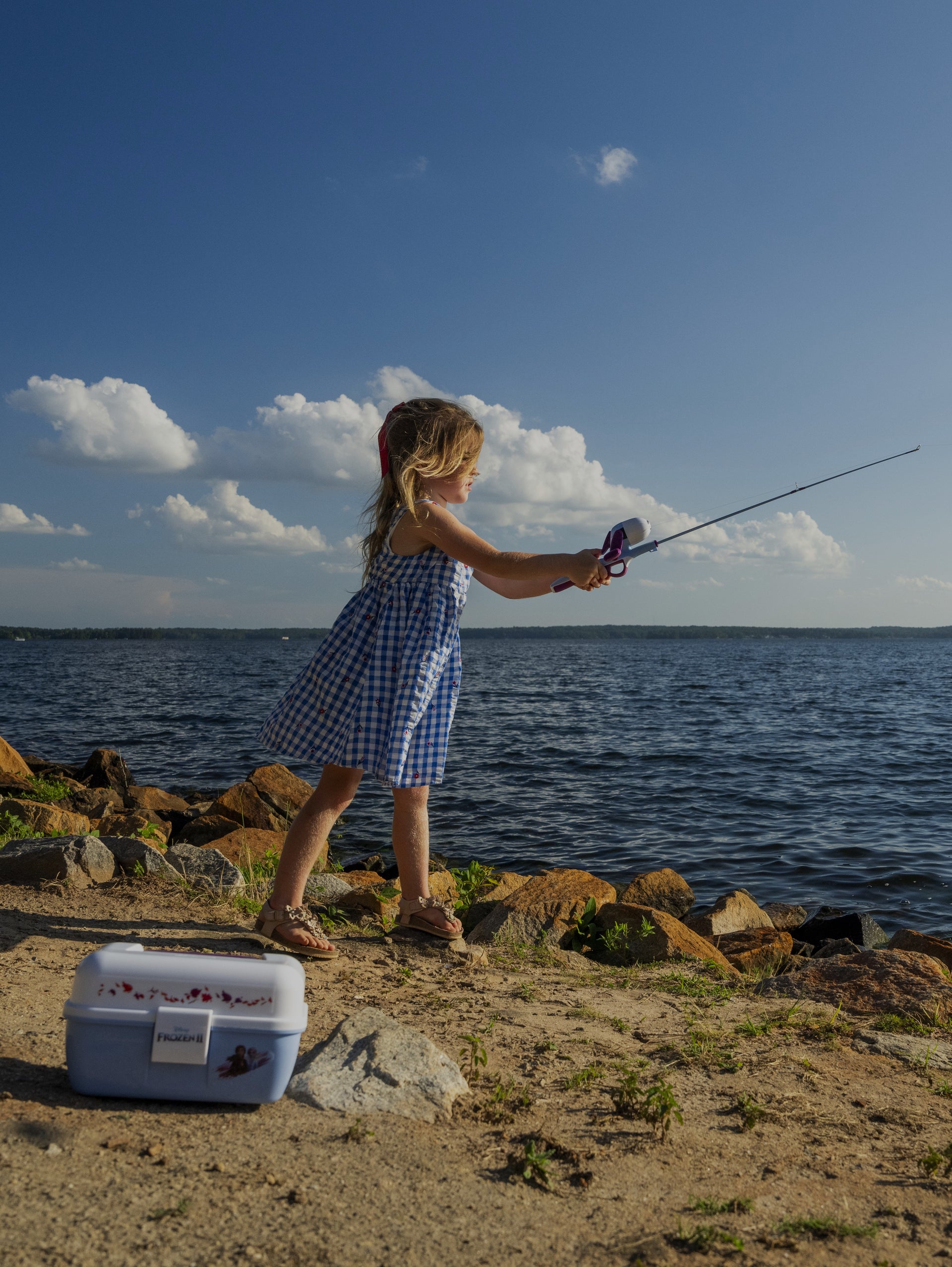 Child in a blue checkered dress fishing by a lake with a cooler nearby.